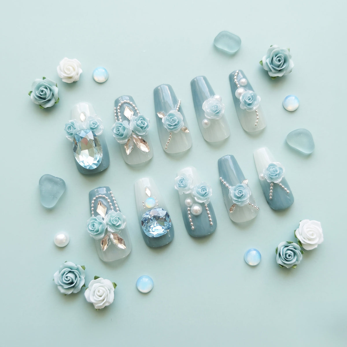 A flat-lay of decorative press-on nails arranged on a pale mint background, with scattered matching flower and bead embellishments. The nails feature soft blue-gray and white gradients with floral details, pearl-like dots, metallic gold accents, and clear rhinestone embellishments.