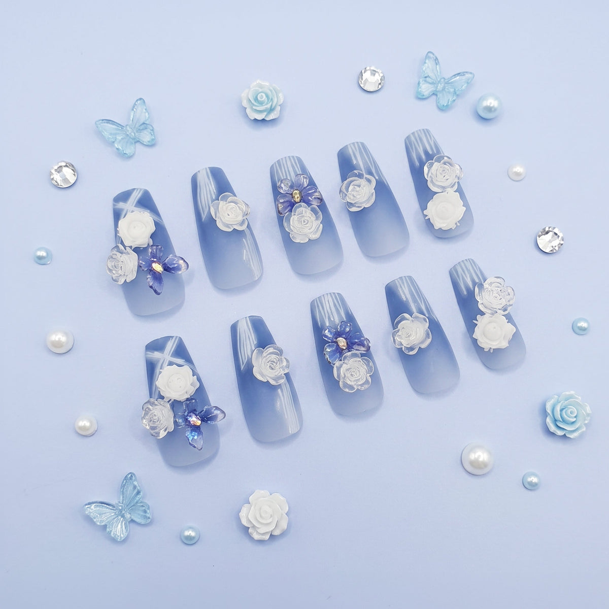 A set of decorative press-on nails arranged on a light blue background, featuring a glossy light blue base with white floral embellishments, white pearl-like accents, and small blue butterfly details.