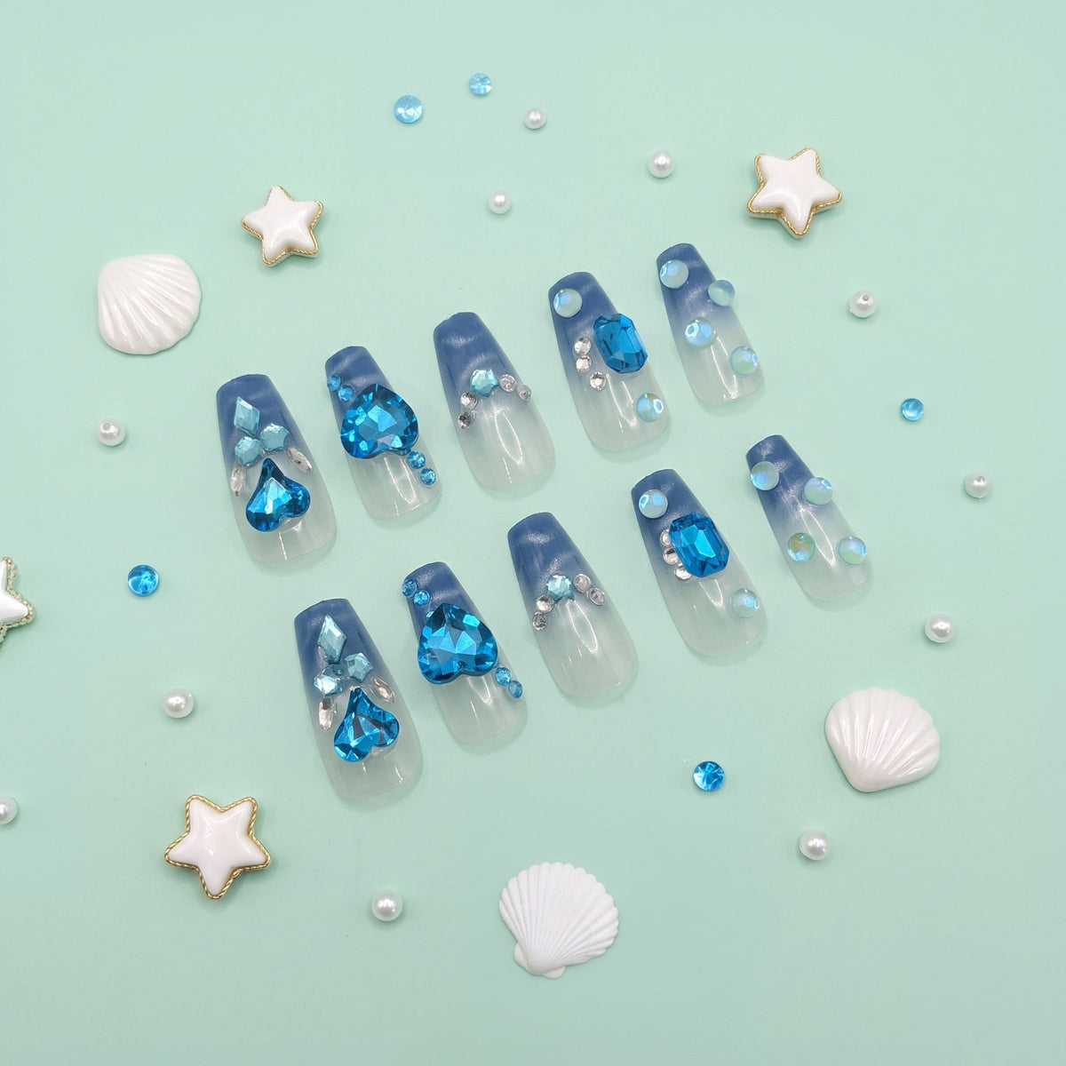 A set of false press-on nails arranged on a mint green background with decorative seashells, starfish, and small blue rhinestones. The nails are clear with a blue gradient top and contain embedded blue and clear rhinestones.